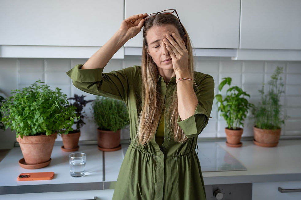 A woman in a green dress standing in a kitchen. She looks fatigued or stressed, holding her glasses in one hand and rubbing her forehead with the other.