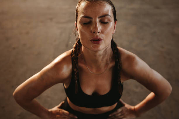 Woman in workout attire, sweating with eyes closed, stands in a dimly lit gym, appearing focused and determined.