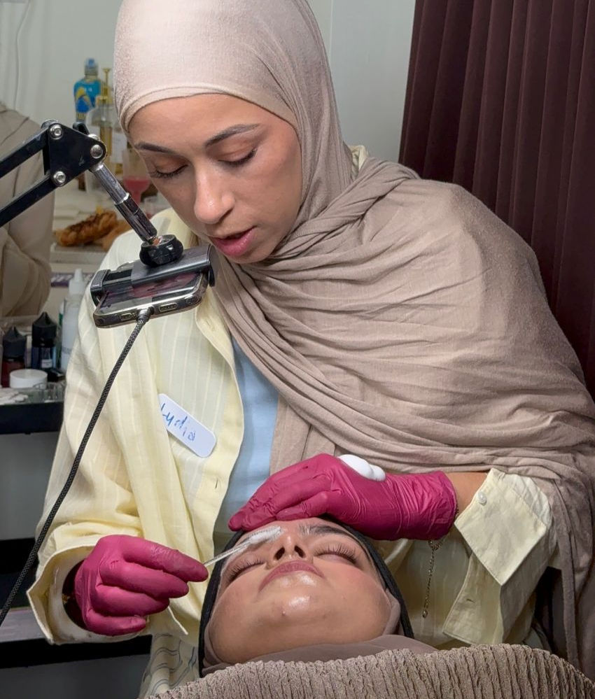 A beautician in a beige hijab and pink gloves applies skincare to a reclining woman's forehead in a salon-like room.