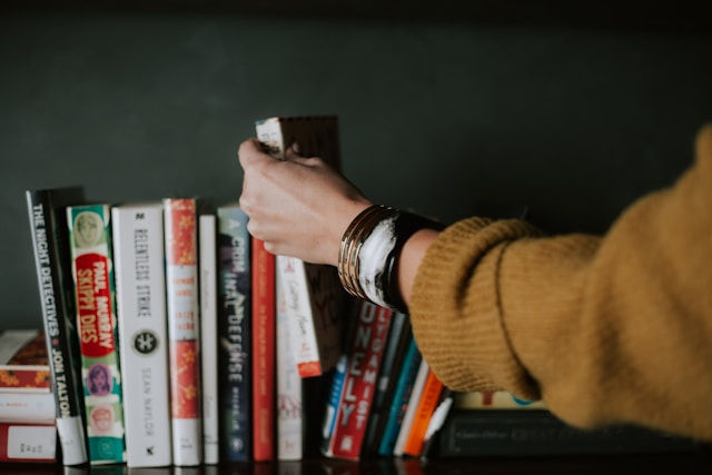 A hand in a yellow sweater reaches for a book on a shelf filled with colorful spines. Text includes book titles like Lonely. Cozy mood.