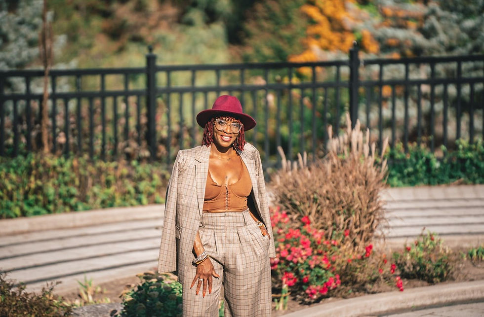 Woman in plaid suit and burgundy hat smiles in a garden with vibrant flowers and black fence, exuding a confident, cheerful vibe.