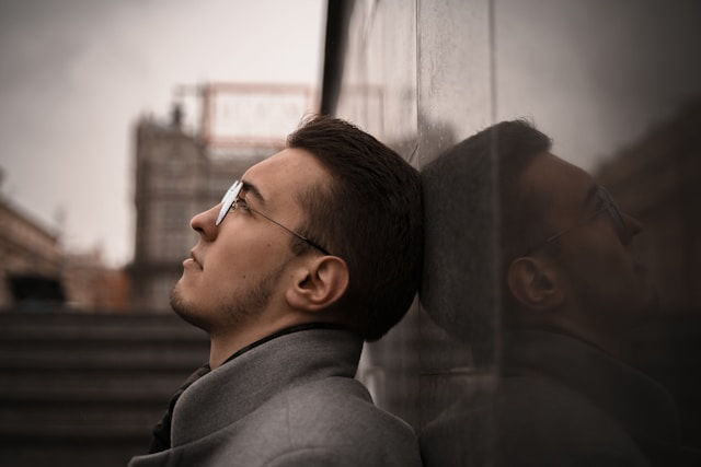 Man in a gray coat leans back against a reflective wall, looking thoughtful. Urban background with a soft, muted sky.