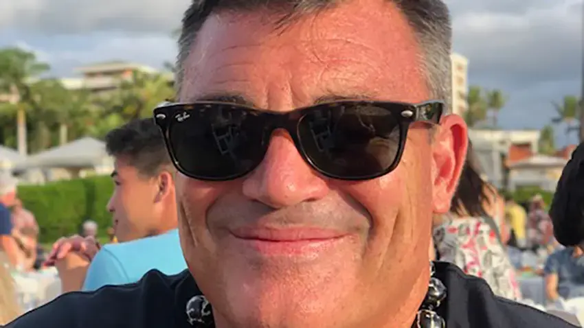 Man wearing sunglasses smiles at an outdoor event. Background shows people, greenery, and a partly cloudy sky, creating a cheerful atmosphere.