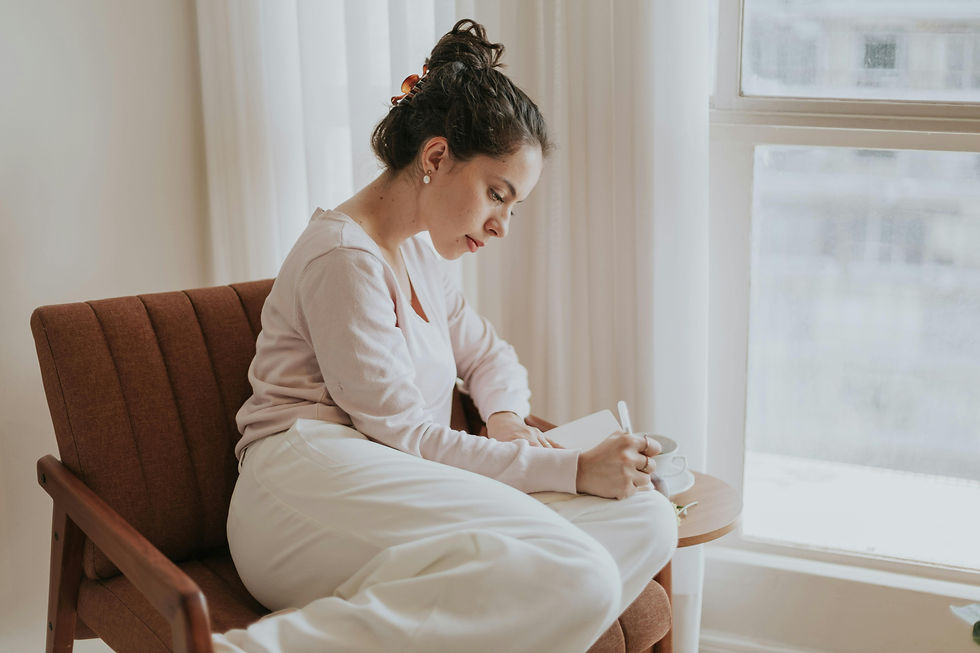 Woman writing in a notebook while seated on a brown chair, next to a window with sheer curtains. She's wearing a light pink top and white pants.