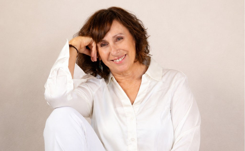 Smiling middle-aged woman in a white shirt sits against a beige wall, resting her head on her hand.