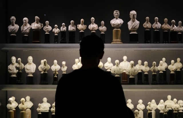 A silhouette of a person observes rows of white classical busts in a dimly lit museum display. The scene feels contemplative and serene.