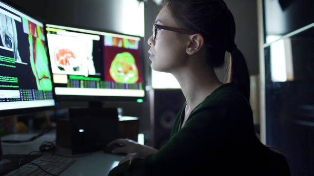 Woman in glasses intently analyzes medical scans on dual monitors in dimly lit room, screen displays bright colors and data.