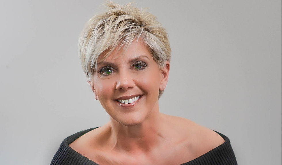Smiling short-haired blonde woman in a black off-shoulder top against a plain gray background.