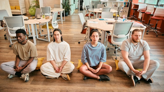 Four people sit cross-legged meditating on a wooden floor in a bright coworking office, calm and focused.