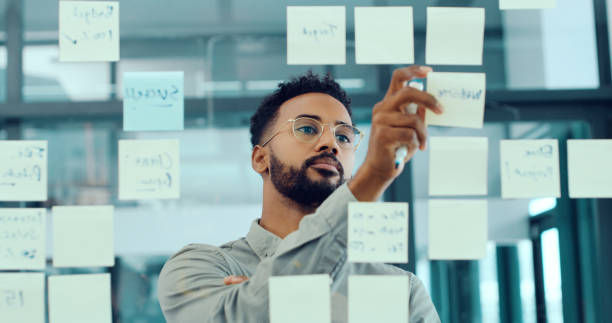 Man in glasses writing on a glass wall covered with sticky notes in an office. Focused expression, blue and white tones, modern setting.
