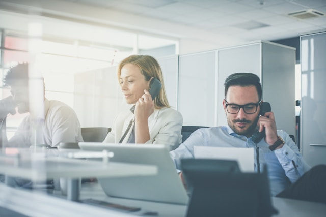 People in an office talking on phones, using laptops. Bright lighting, modern workspace. Professional, focused mood.