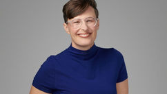 Smiling person with short hair and glasses wearing a blue top against a gray background. They convey a friendly and approachable mood.