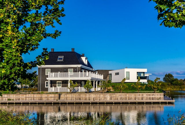 The photo showcases two modern waterfront homes, one black with white accents and a wraparound balcony, and the other sleek and white, both reflecting beautifully on a calm waterway.