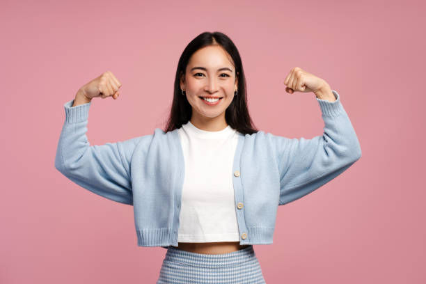Smiling woman in a blue cardigan flexes her arms confidently against a pink background. She wears a white top and a blue skirt.