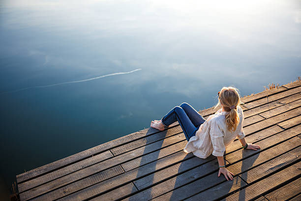 A person sits on a wooden dock, gazing at calm blue water. The setting is peaceful with warm light. They're wearing a white top and jeans.