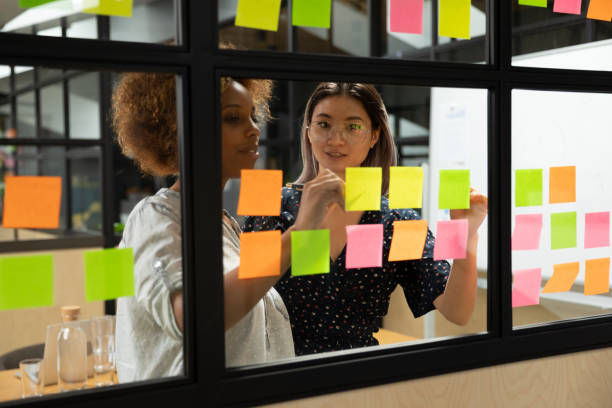 Two women in an office collaborate, arranging colorful sticky notes on a glass wall. They appear focused and engaged in their task.