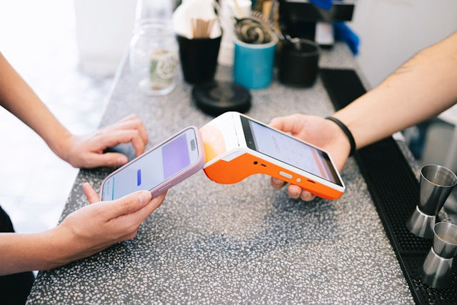 Two people complete a contactless card payment at a counter with a smartphone and orange payment terminal