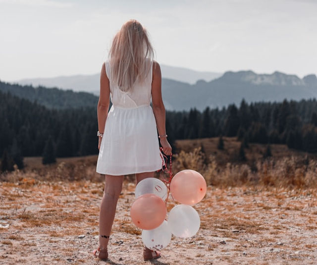 The photo shows a woman standing outdoors with her back to the camera, holding pink and white balloons.