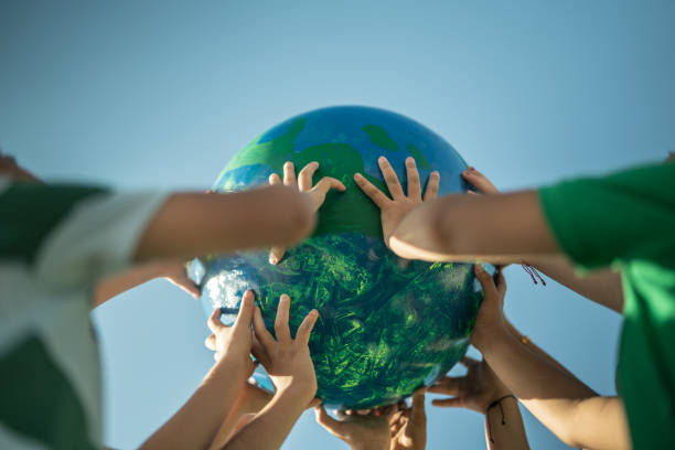 Hands lifting a globe against a clear blue sky. The globe is blue and green, symbolizing Earth. People wear green shirts, conveying unity.