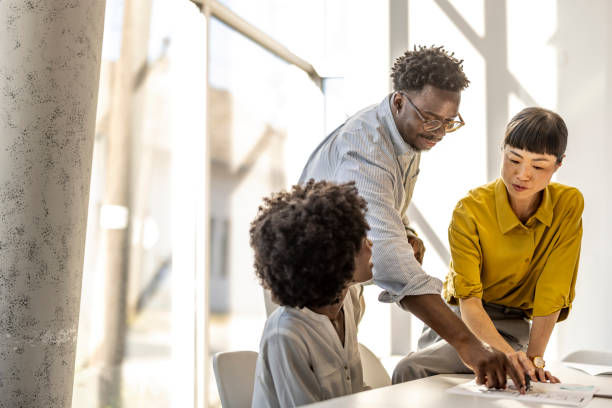 Three colleagues are gathered around a table in a bright office, actively discussing documents and collaborating on a project.