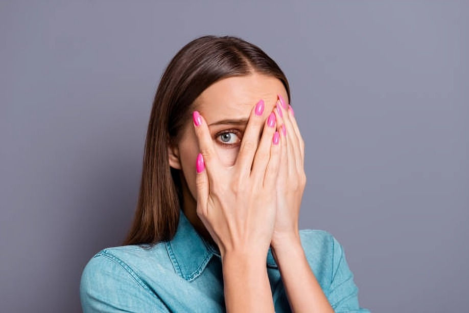 Woman in denim shirt covers face with hands, revealing one eye. She has pink nails and appears anxious. Gray background.