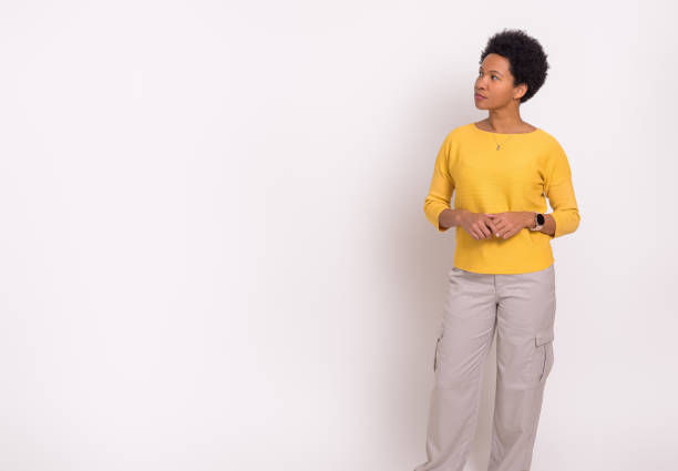 Person in a yellow sweater and beige pants stands against a plain white background, looking thoughtfully to the right.