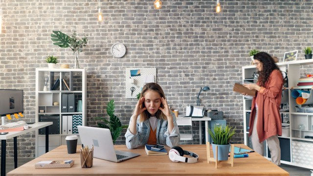 Woman sits at a desk with laptop and headphones, looking stressed, while another woman checks a clipboard in a modern brick office.