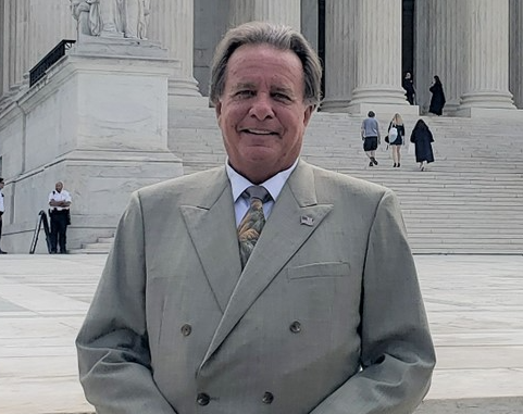 Smiling man in a light gray suit stands on courthouse steps before tall columns and a seated statue, holding a folder.