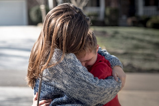 Woman in a gray sweater hugs a young boy in a red shirt outdoors, comforting him in a tender, emotional moment.