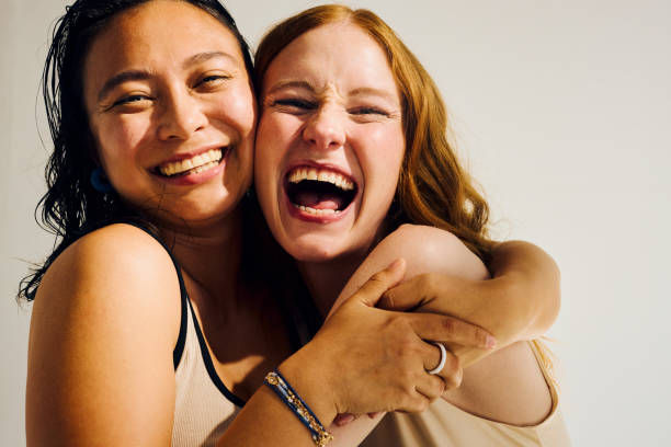 Two women joyfully embrace, laughing together against a plain background. They wear neutral-toned tops and appear to be in a cheerful mood.
