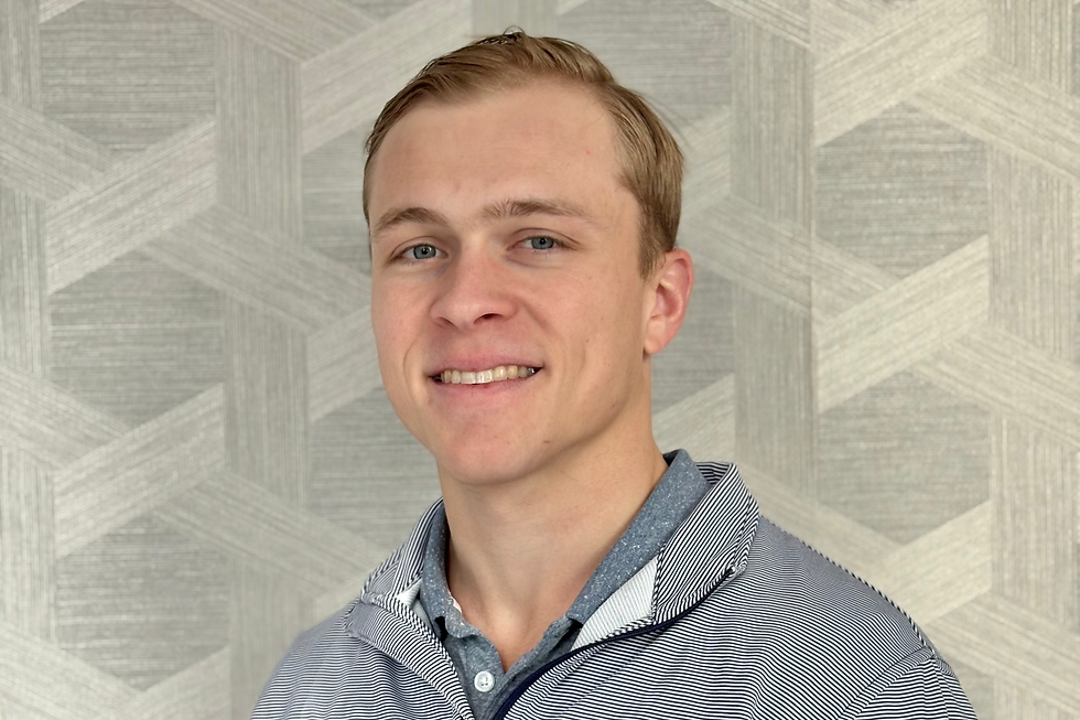 Young man smiling in front of a gray geometric-patterned wall, wearing a striped pullover and blue shirt, conveying a friendly mood.