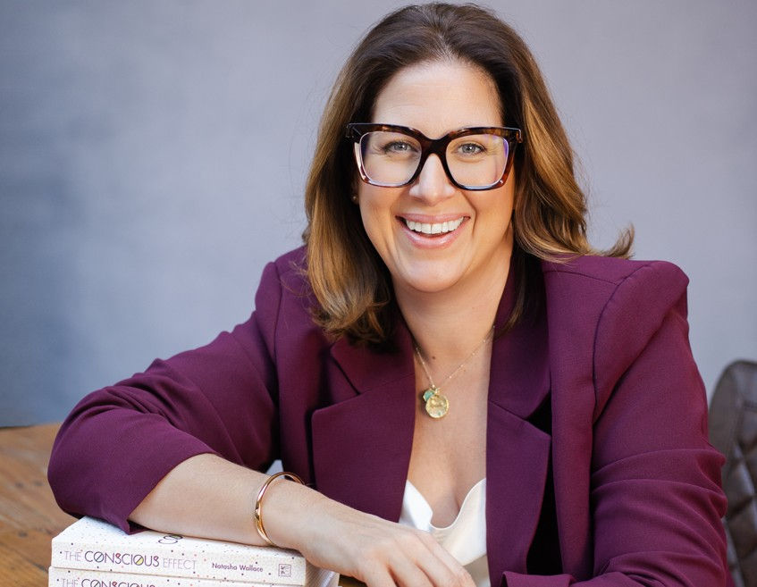 Smiling woman in glasses and a purple blazer leans on books, including The Conscious Effect, against a gray wall.