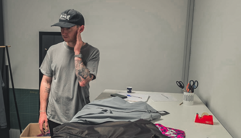 Man in a gray shirt and cap stands by a worktable, touching his face, with fabric, scissors, and papers in a calm studio.