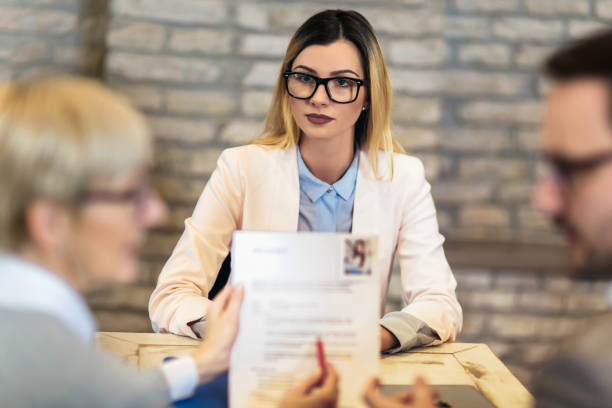 The photo shows a young woman with long blonde hair, wearing black-rimmed glasses and a light-colored blazer, sitting across from two blurred interviewers in a professional setting.