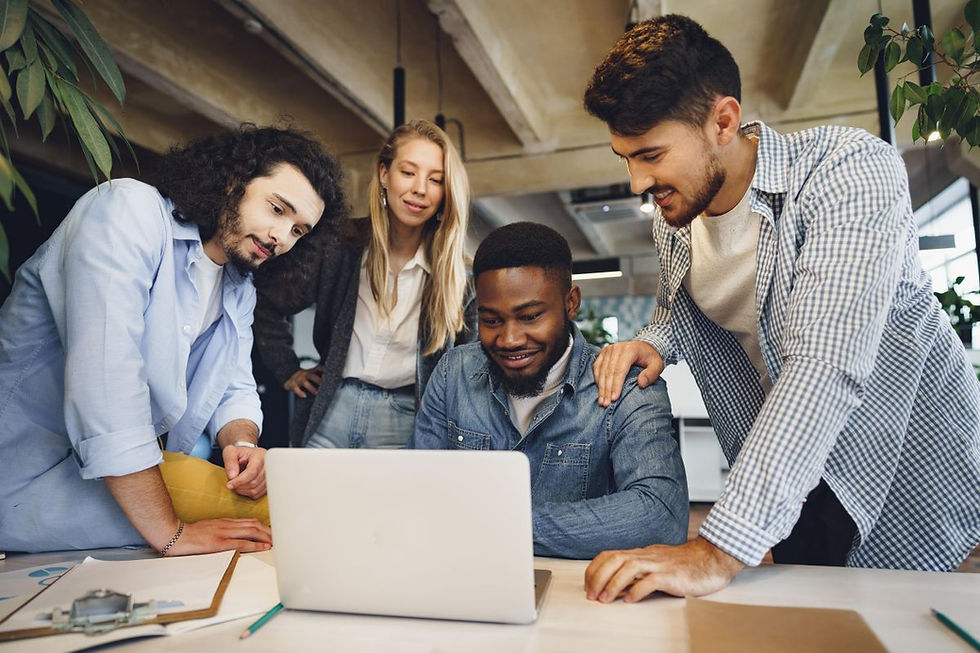 Four coworkers smile around a laptop in a modern office, leaning in during a team discussion.