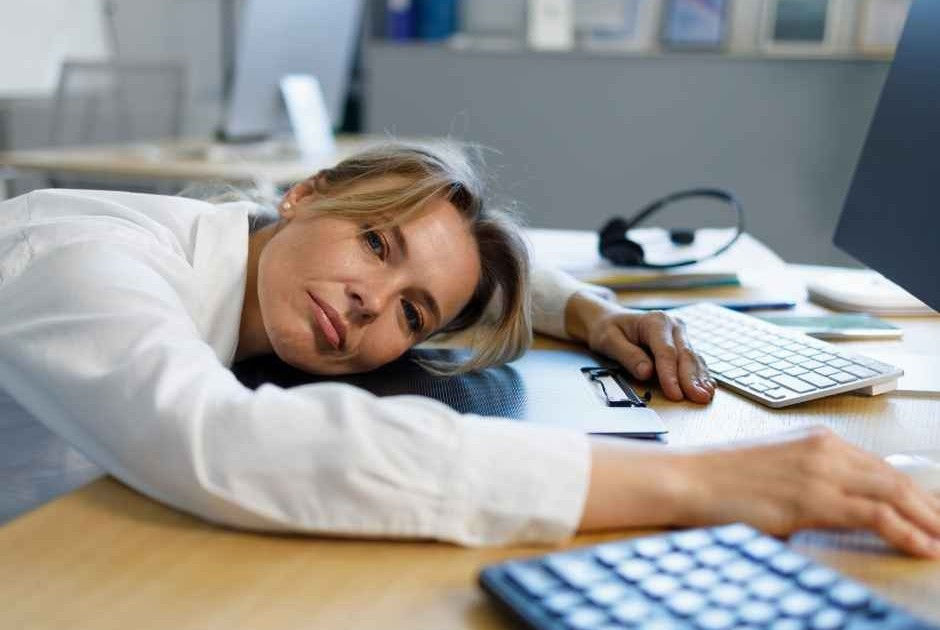 Blonde office worker rests her head on a desk, reaching toward a keyboard, looking tired in a bright office.