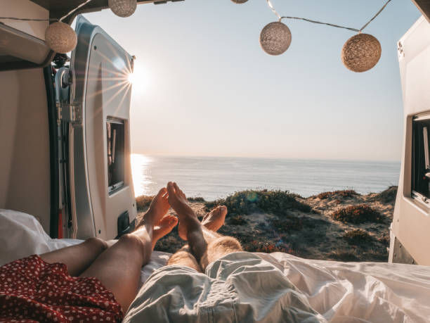 Two people relax in a van by the sea, legs outstretched towards the sunset. Warm tones and string lights create a serene atmosphere.