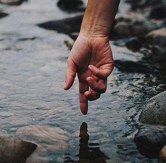 A hand gently touches clear water amidst smooth stones in a creek, evoking a serene and tranquil mood.