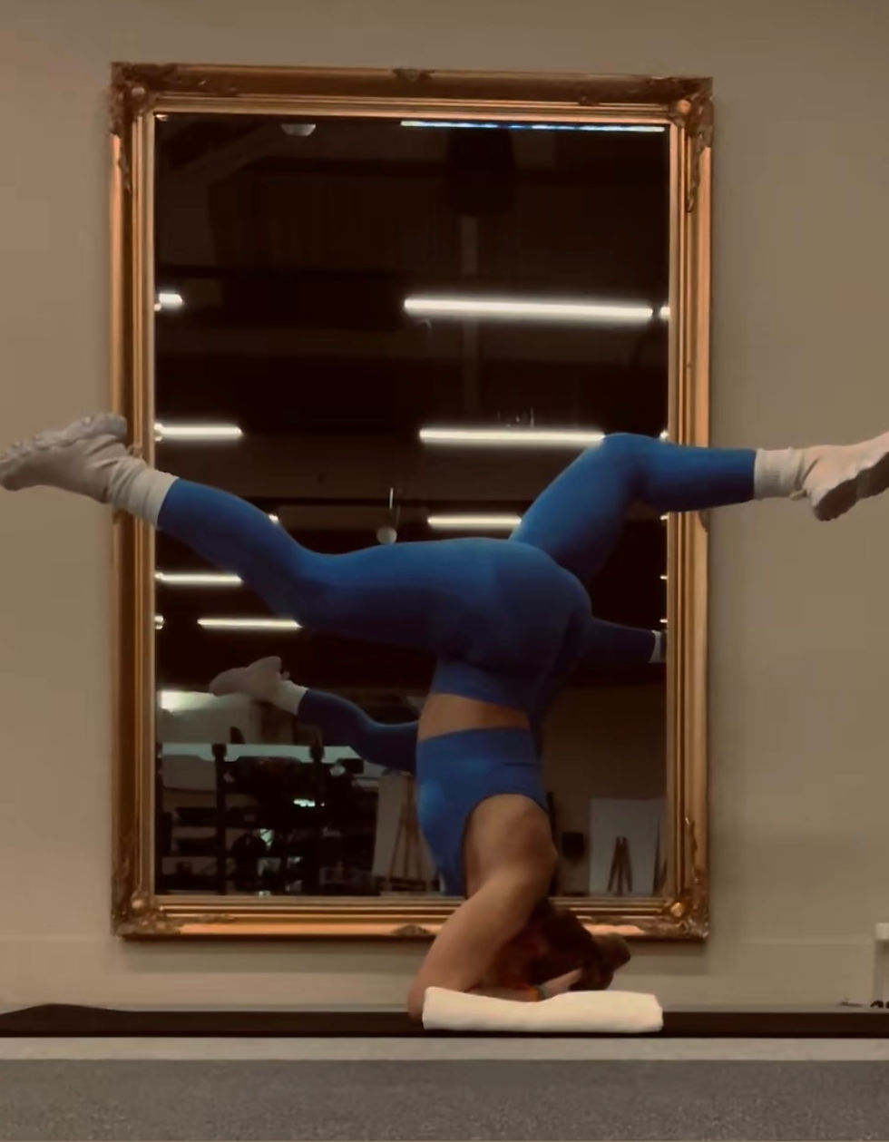 A person in blue activewear performs a headstand in a gym, reflected in a large mirror. The setting is calm with fluorescent lights.