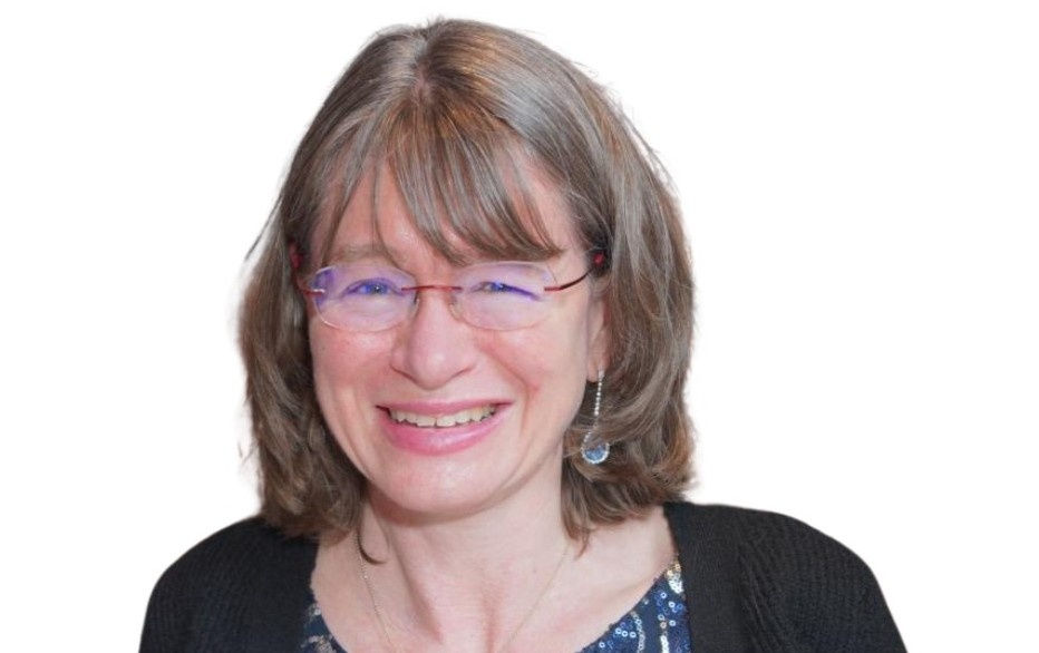 Smiling woman with glasses, gray hair, wearing a black cardigan and blue patterned top. White background. Mood: cheerful.
