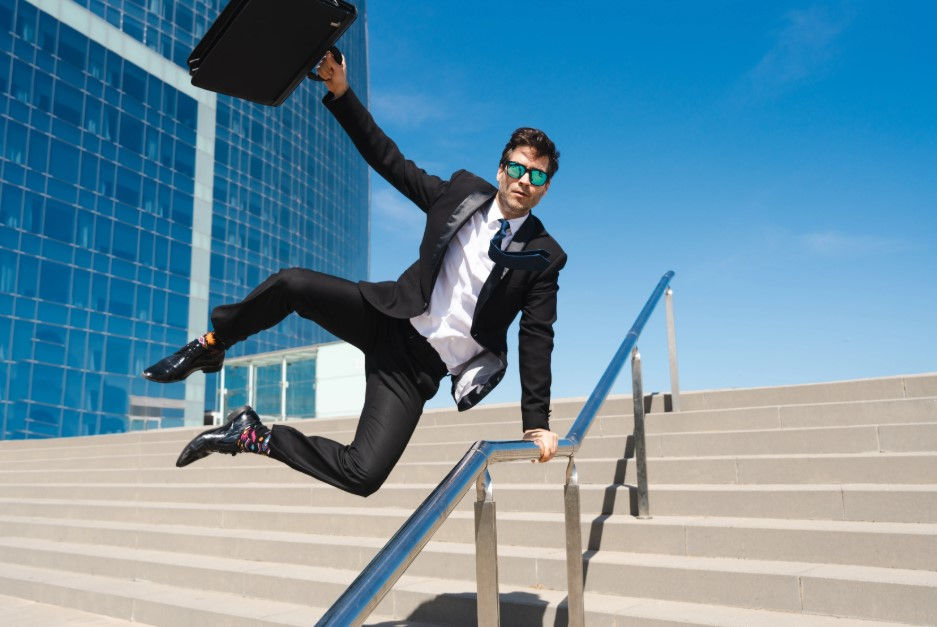 A man in a suit, holding a briefcase, energetically jumps over a railing on outdoor stairs, with a modern glass building and blue sky in the background.