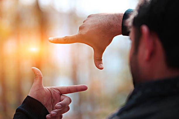 A man is framing a scene with his hands, creating a rectangle shape with his fingers while looking toward a glowing light in the blurred background.