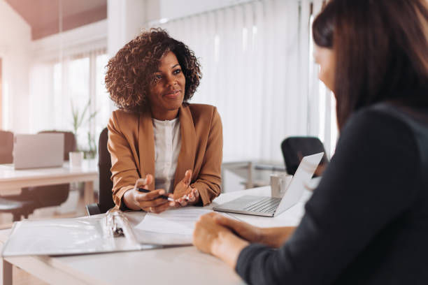 Two women in a bright office meeting, one speaking with a phone and papers beside an open laptop.