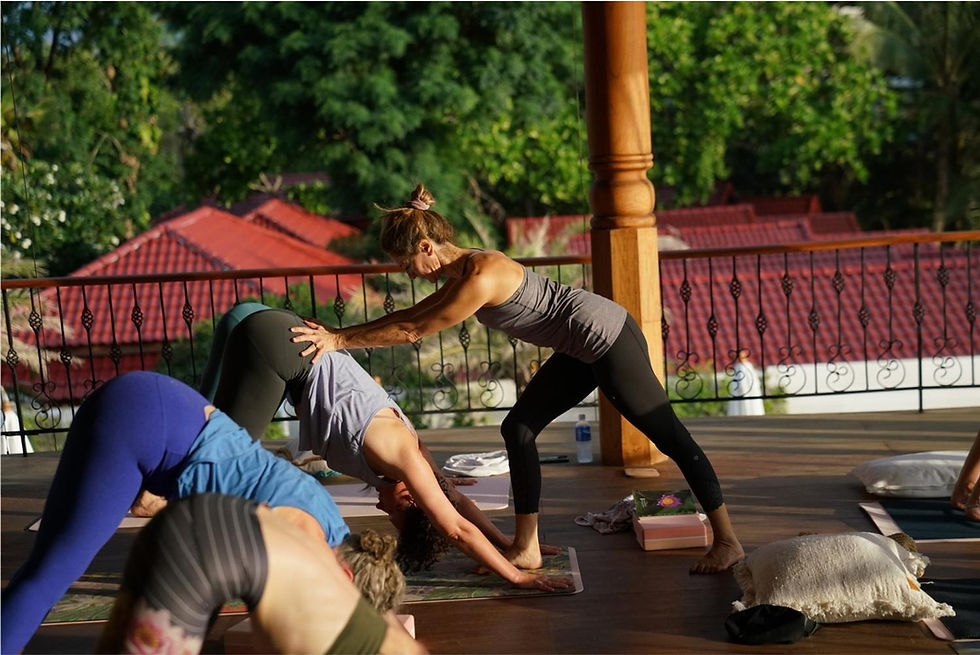 Yoga instructor assists a person in a downward dog pose on a sunny wooden patio with red rooftops and greenery in the background.