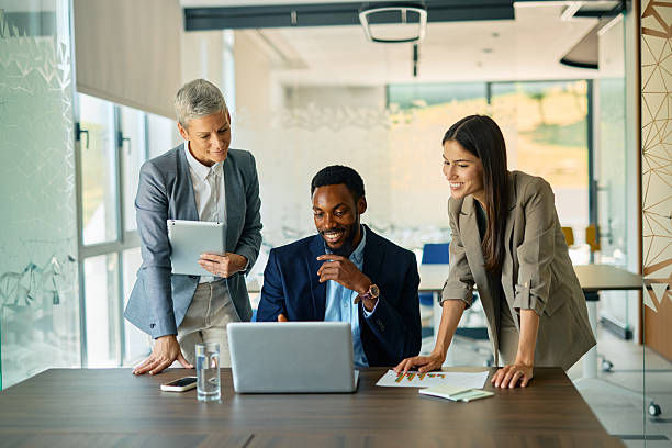 Three colleagues collaborate happily around a laptop in a modern office, one holding a tablet. Bright setting and professional attire.