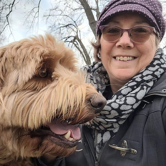 Woman with glasses and a purple hat smiles beside a fluffy brown dog. She's wearing a gray patterned scarf. Trees are in the background.
