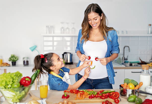 A woman and a young girl are in a bright kitchen preparing fresh vegetables. The girl is playfully holding pieces of bell pepper and cherry tomatoes near the woman’s belly, making her smile.