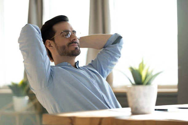 Man in glasses relaxing with hands behind his head, sitting at a desk with potted plants, in a bright room with large windows.