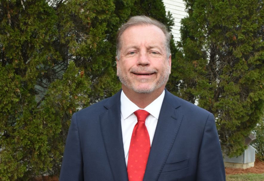 Man in a navy suit and red tie smiling in front of lush green trees. The mood is formal and positive. No text visible.