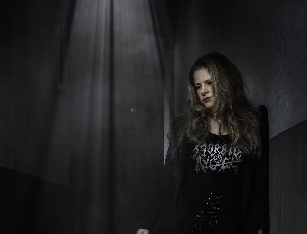 Woman with long hair in a black outfit leans against a dark wall, illuminated by a beam of light. Her shirt has the words "Morbid Angel."